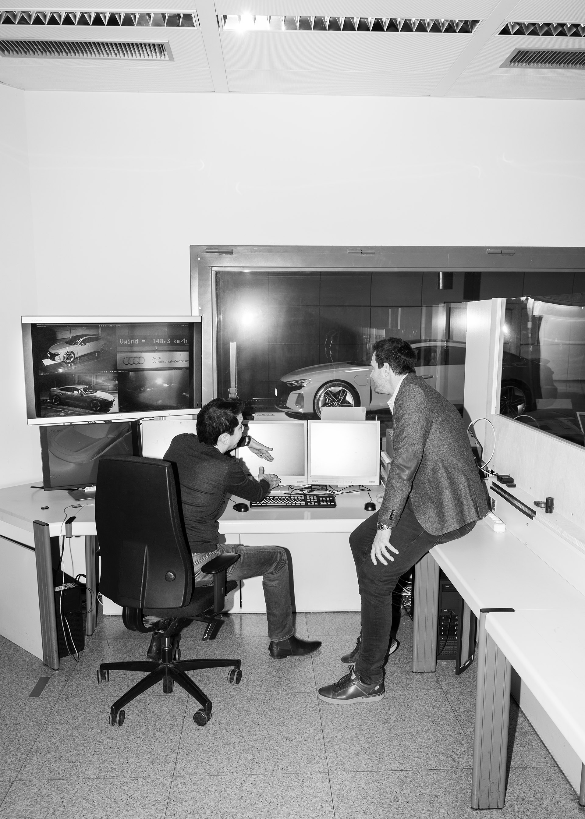 Dr. Kentaro Zens und Thomas Redenbach im Gespräch vor mehreren Bildschirmen, im Hintergrund hinter einer Scheibe der Audi RS e-tron GT.